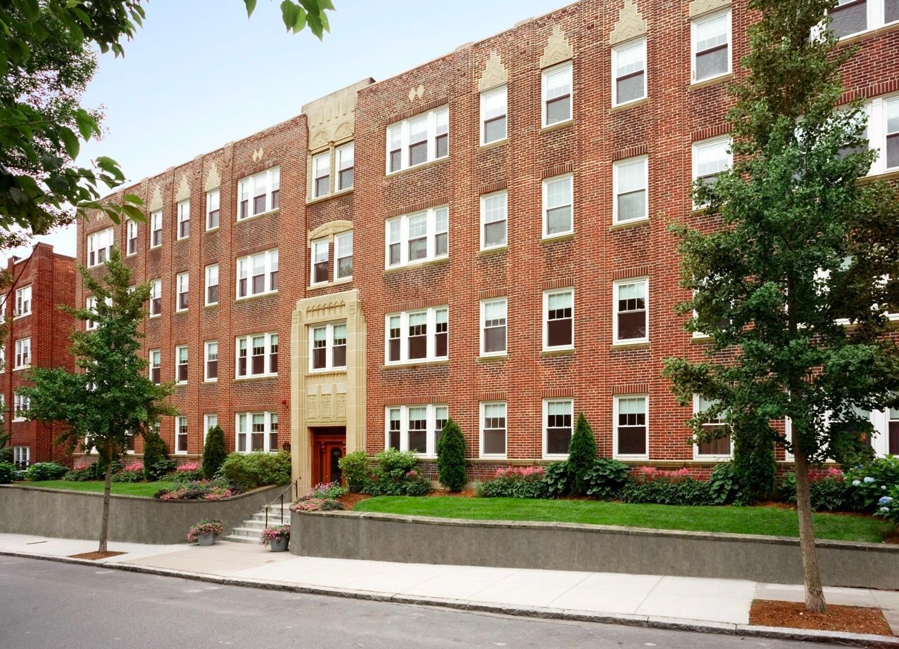 Brick apartment building exterior with entrance, trees, and landscaped front yard.