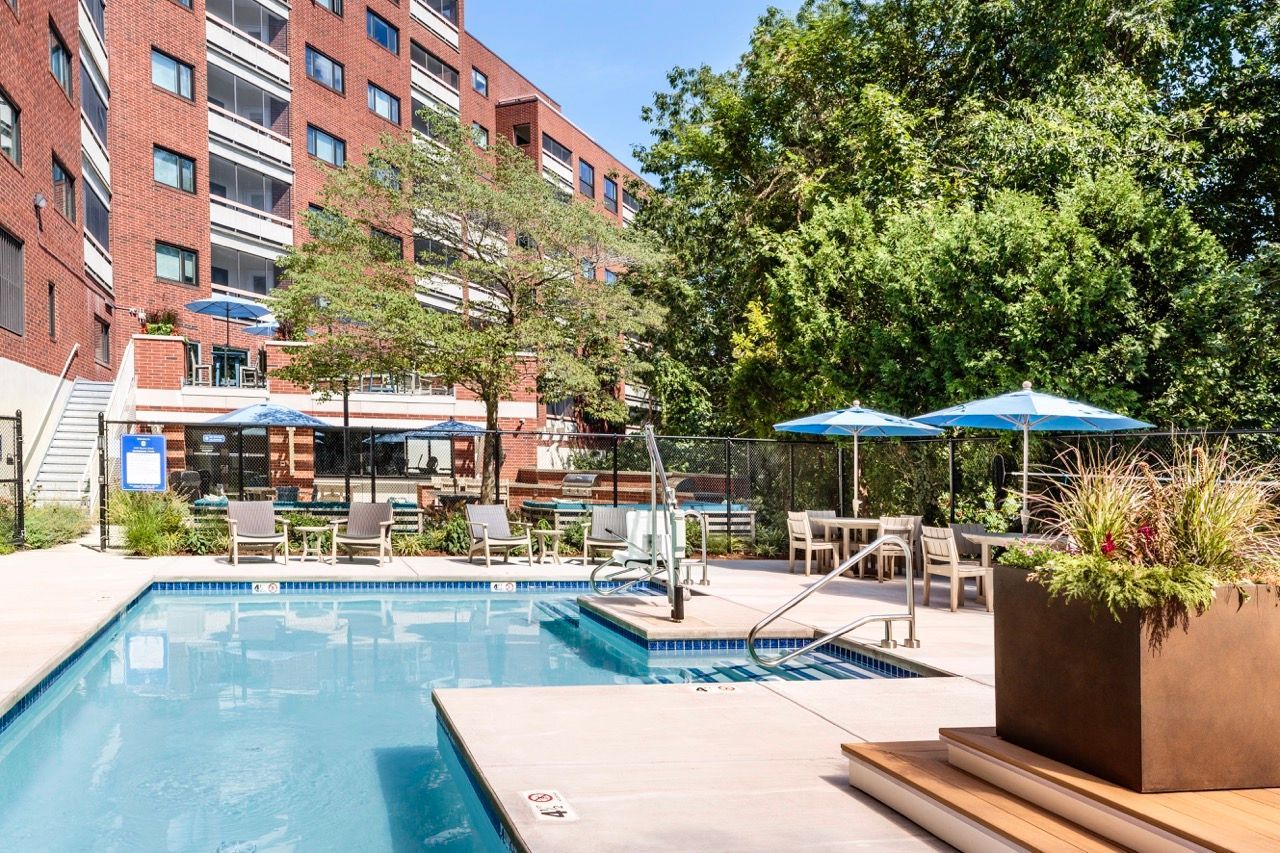 Outdoor community pool area with lounge chairs, blue umbrellas, and landscaping.