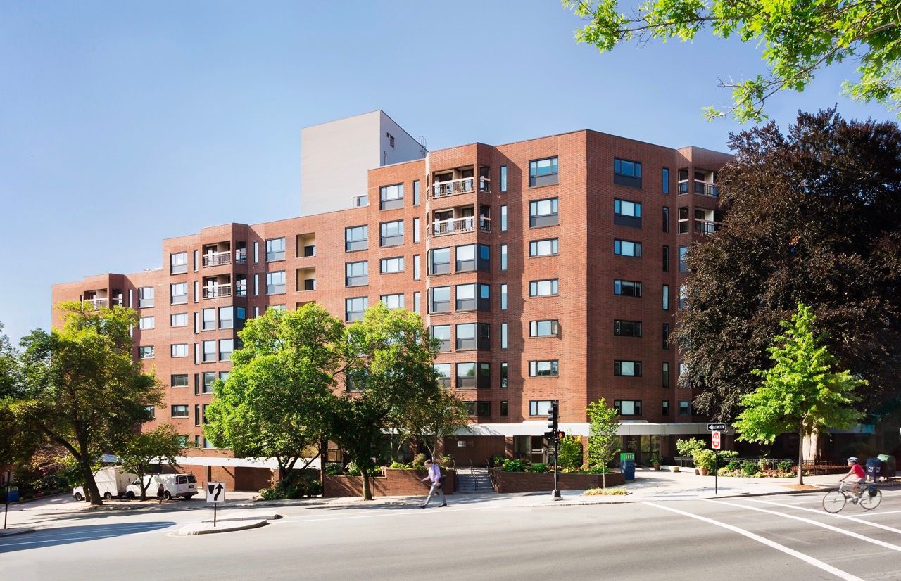 A multi-story brick apartment building on a sunny day with trees in the foreground and a street intersection nearby.