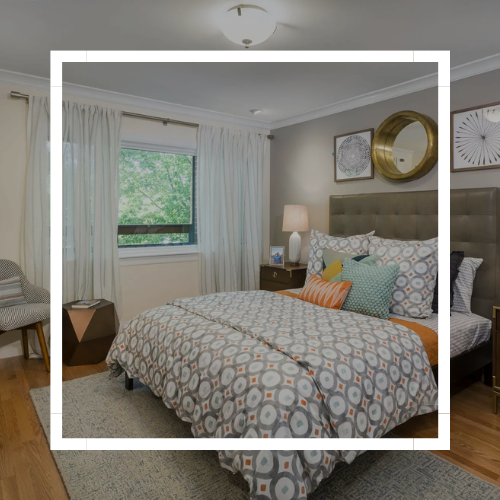 Neatly made bedroom with patterned bedspread, white curtains, bedside lamp, and wall art by a window.