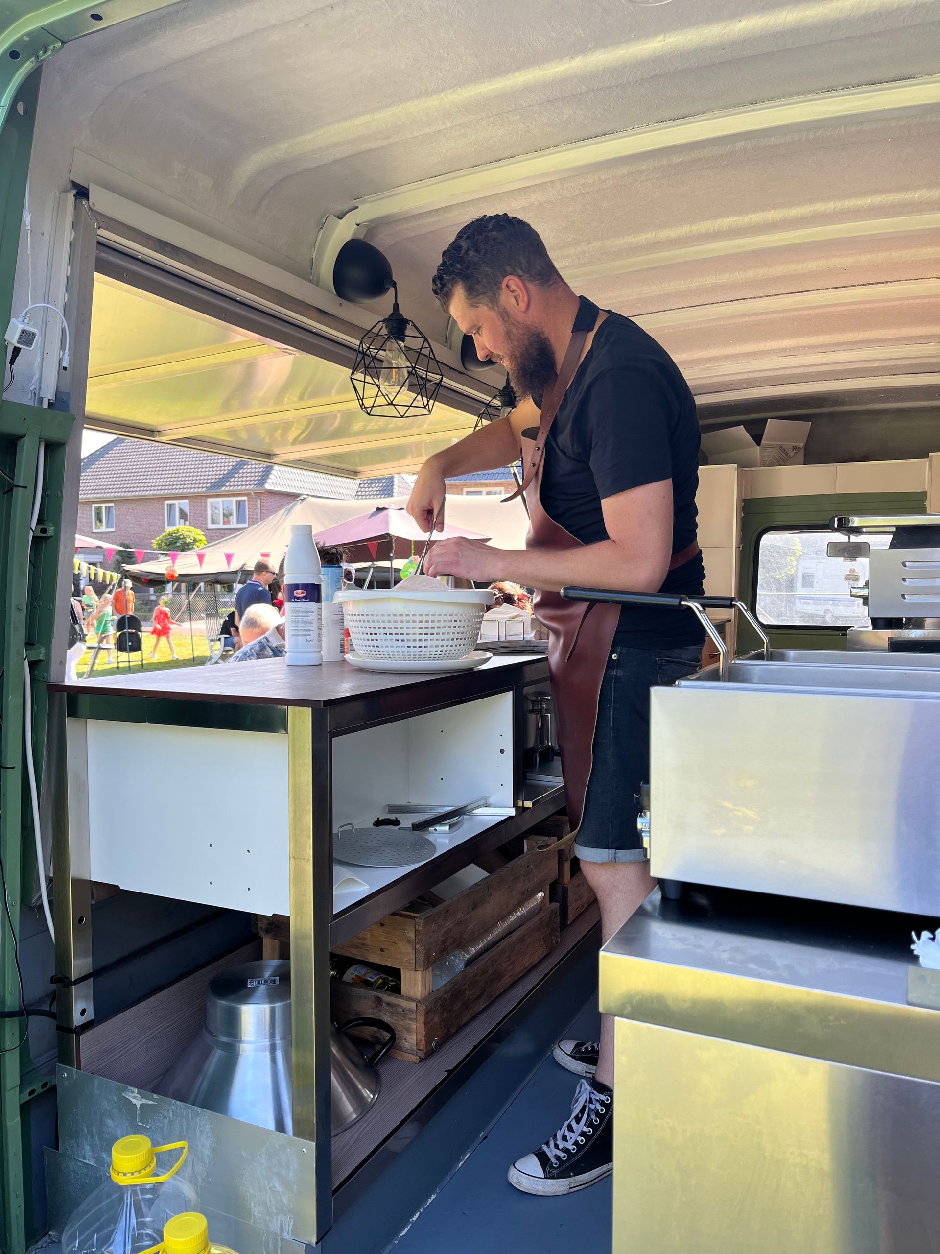 Een foodtruckeigenaar in een zwart T-shirt en een bruin schort werkt in een foodtruck en bereidt eten in een witte kom.