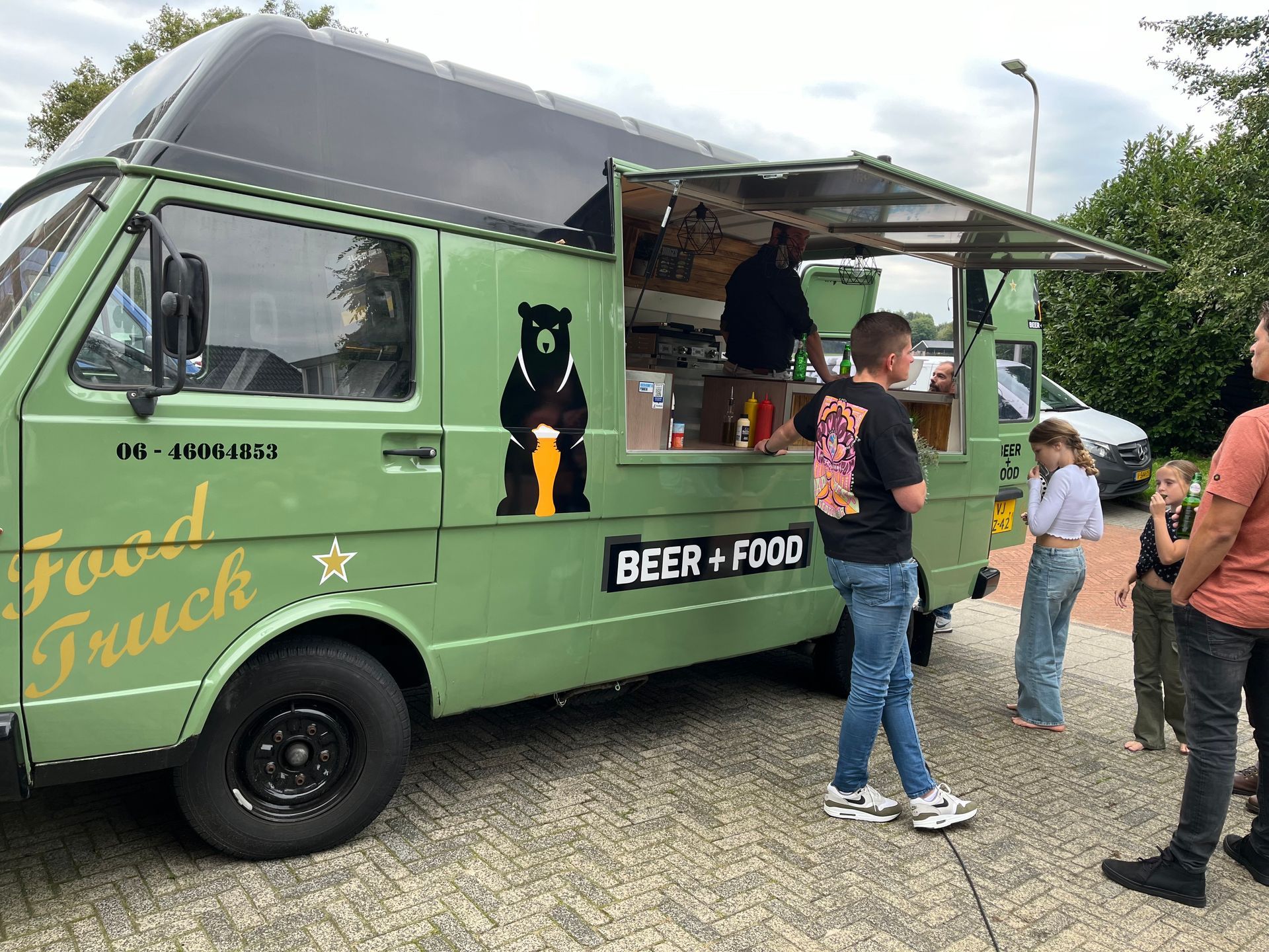 Een lichtgroene foodtruck met bier en eten, voorzien van een berenlogo, staat buiten geparkeerd en bedient klanten via een open zijraam.