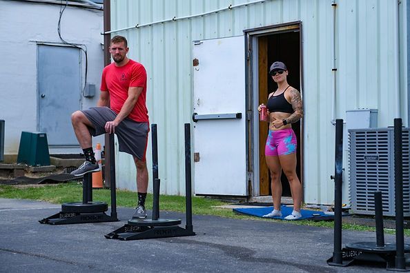Two people in workout clothes on a gym sled track outside CrossFit Bohica in Goldsboro, NC, one leaning on a sled platform and one standing in a doorway