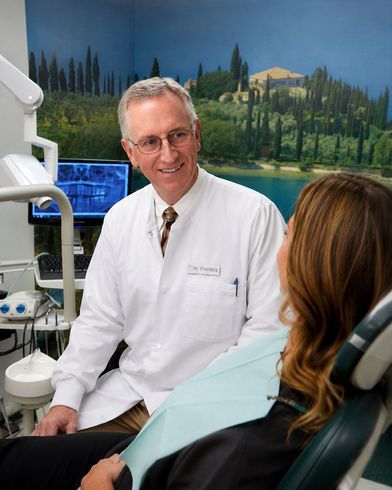 Dentist in white coat speaking with seated patient in a dental exam room