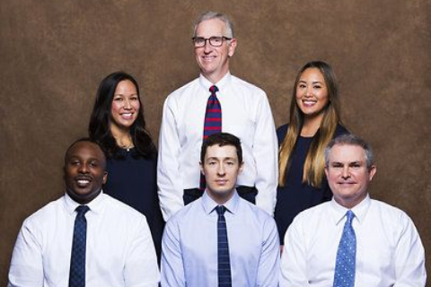Six people in business attire posing for a formal group portrait against a brown backdrop.