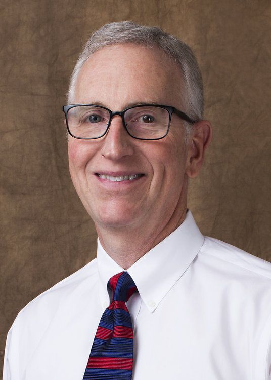 Portrait of a smiling man in glasses, white shirt, and red-blue striped tie against a brown backdrop