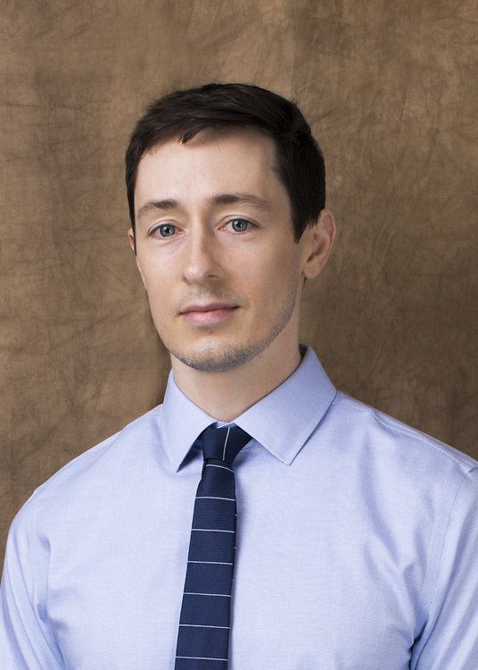 Man in a light blue shirt and striped tie, posed against a plain brown studio backdrop.