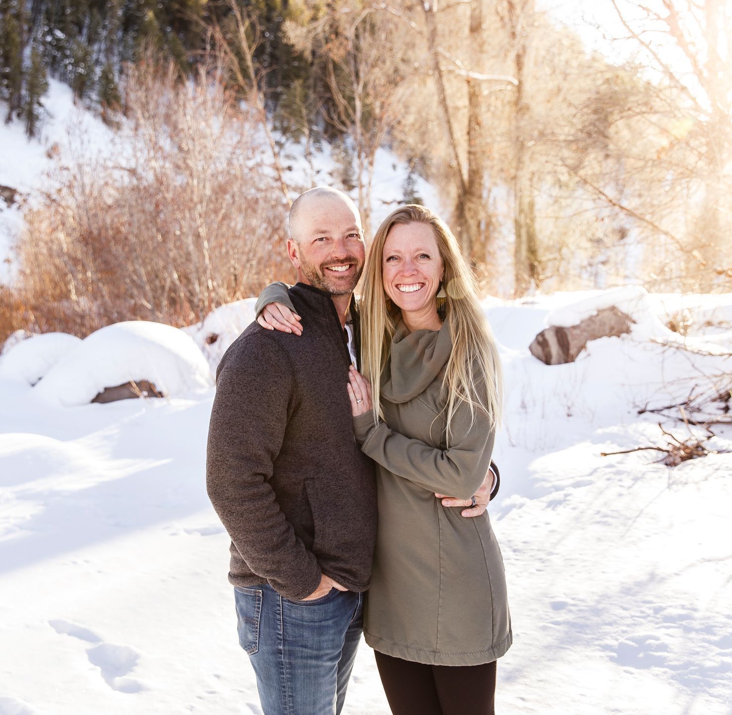 A smiling couple in winter clothing poses together in a snowy, sunlit outdoor setting.