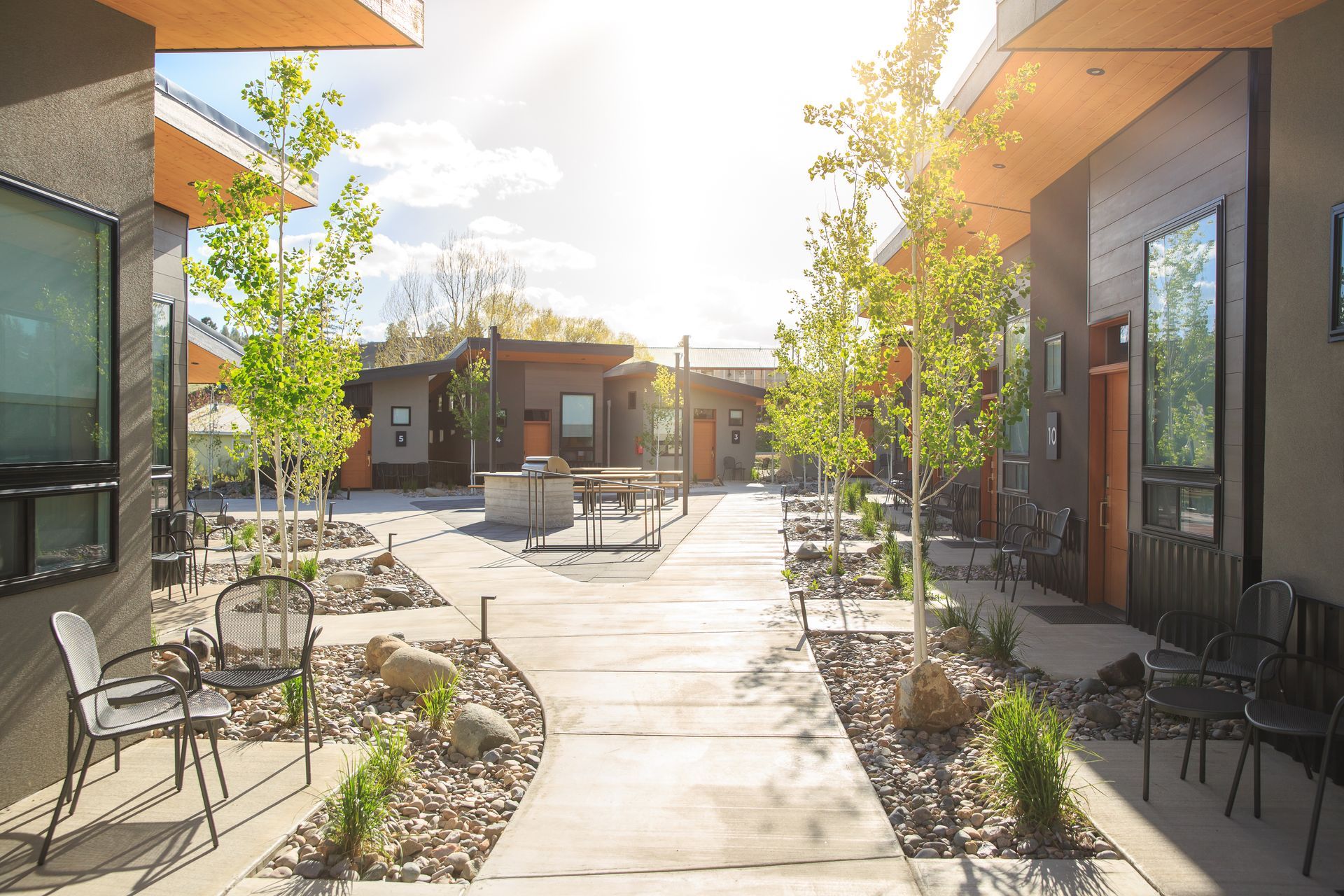 Modern courtyard with a central concrete walkway, surrounded by dark-toned single-story dwellings, chairs, and trees.