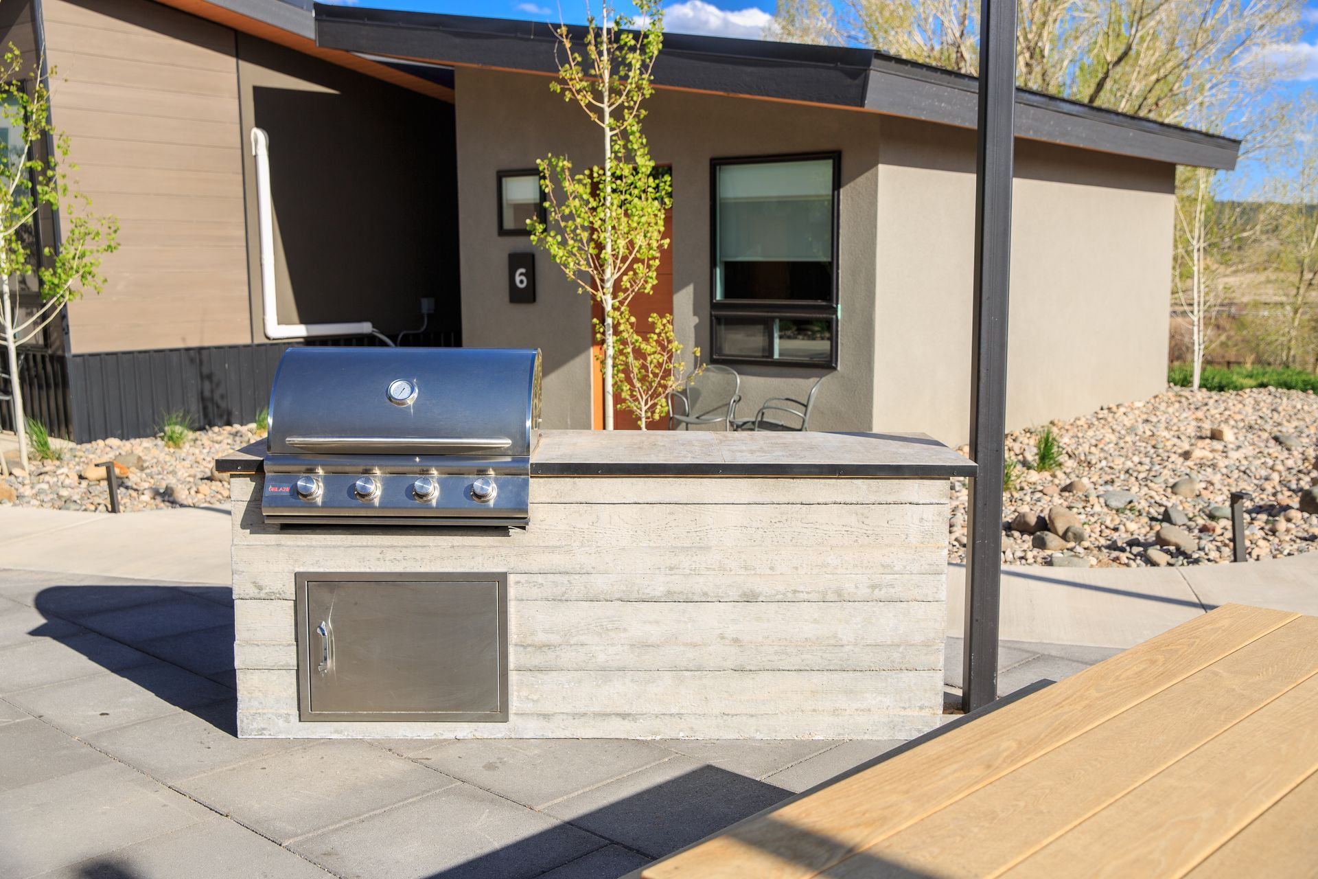 Outdoor kitchen island with a stainless steel grill and cabinet door on a paved patio in front of a modern house.