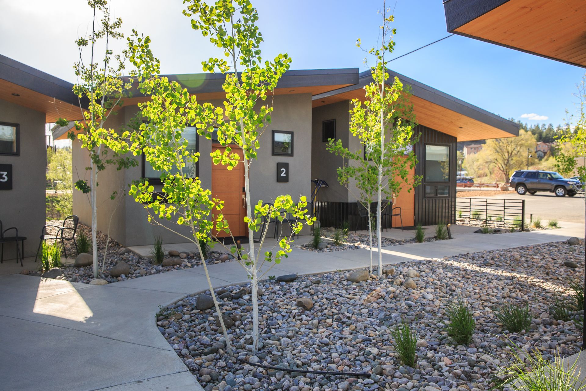 Modern, grey-walled residential units with orange doors and a gravel yard featuring young trees and concrete walkways.