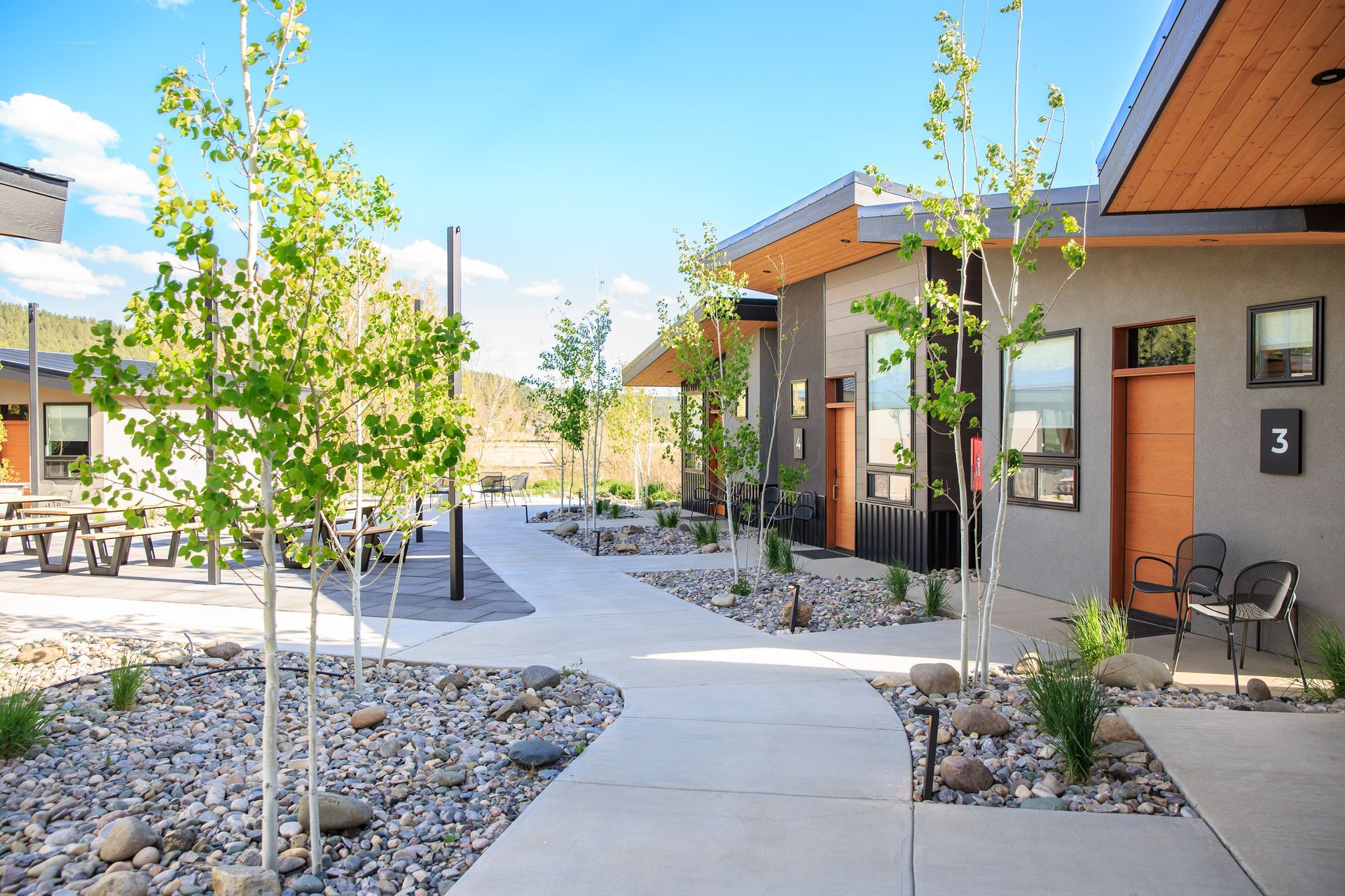 A paved walkway winds between modern buildings with tan exteriors, wooden doors, and gravel landscaping with young trees.