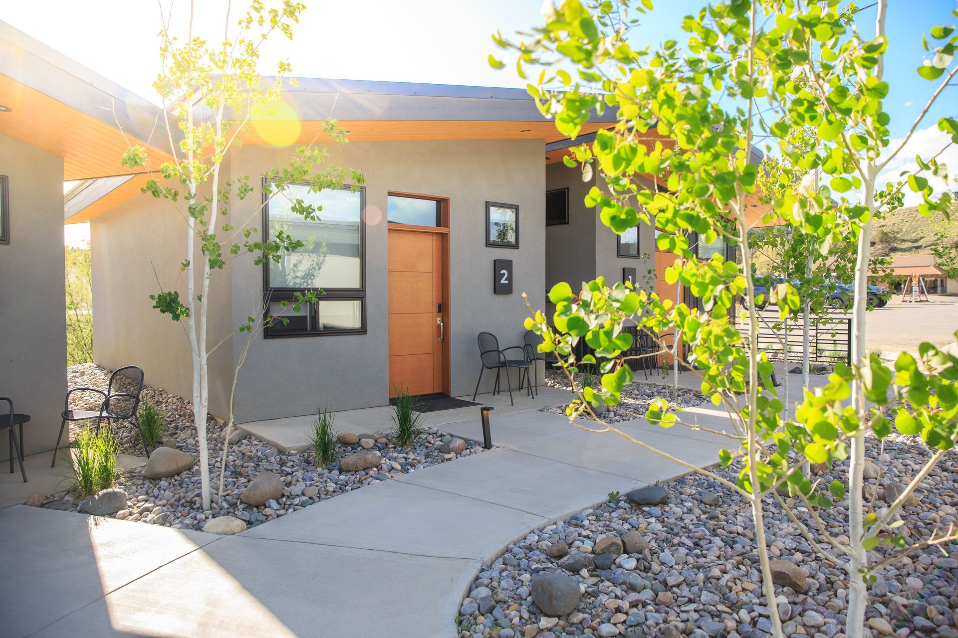 Modern grey buildings with wood doors, a concrete path, and gravel landscaping under a bright sunny sky.