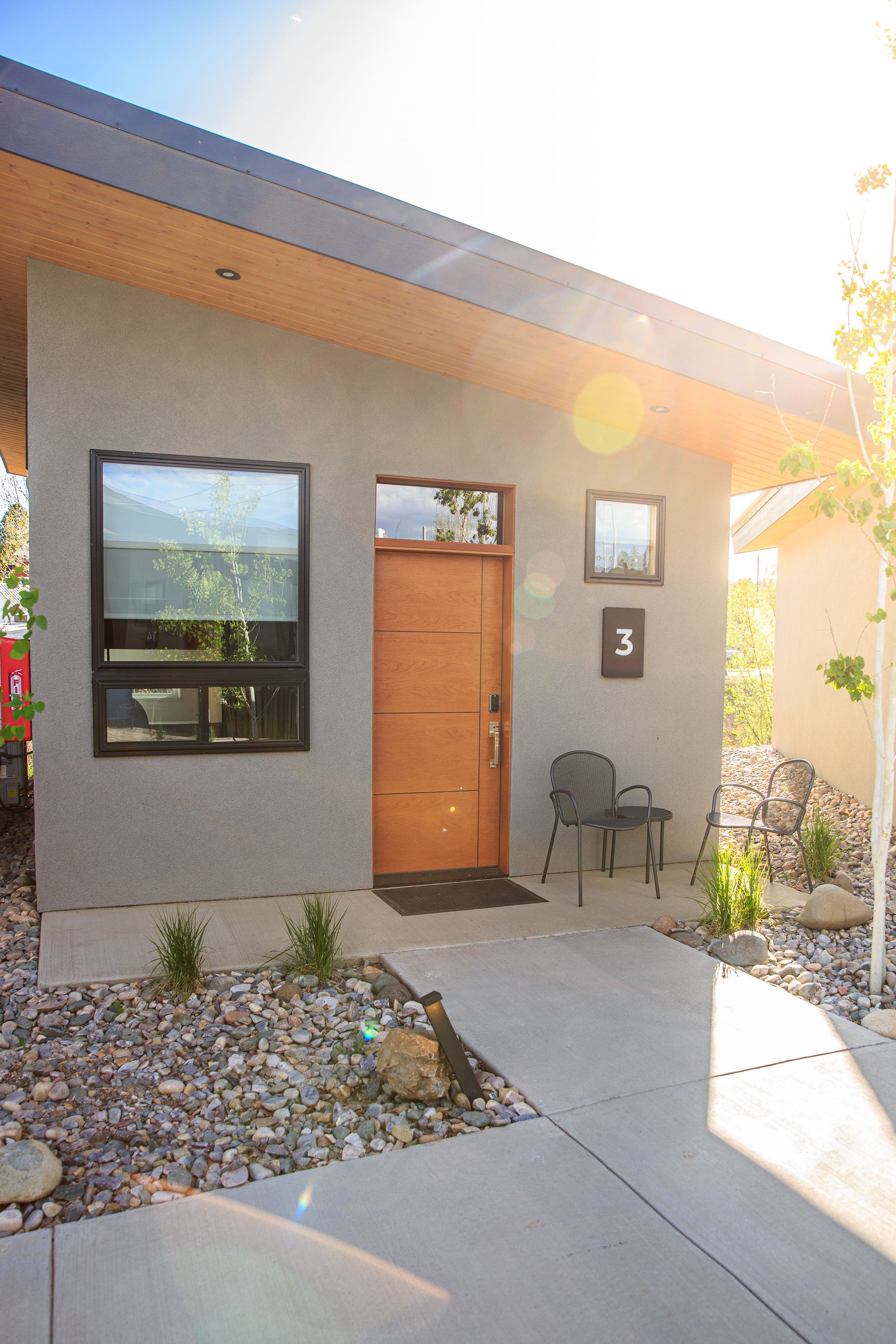 Modern grey house exterior with a wooden door, number 3, and two patio chairs, bathed in warm sunset light.