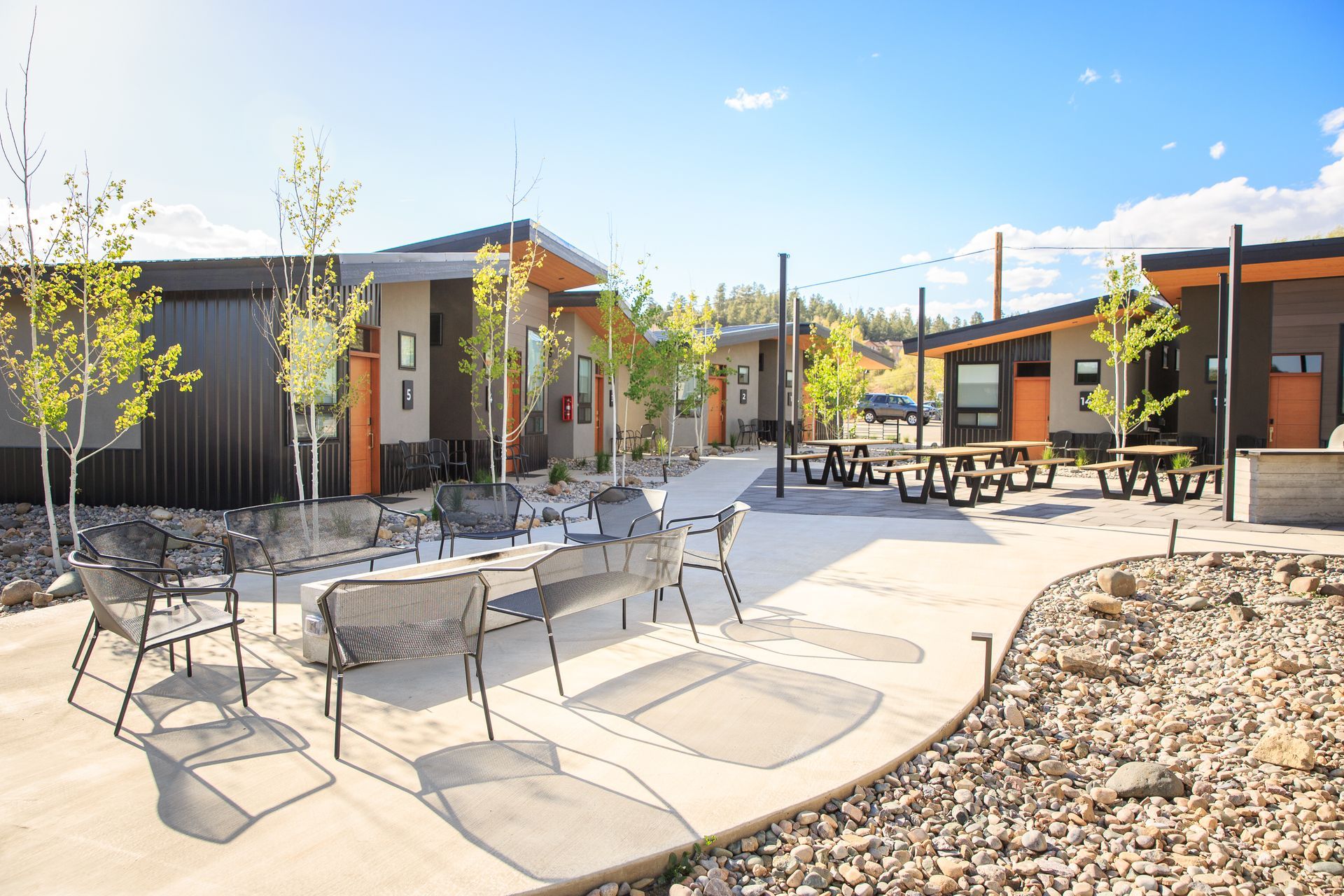 An outdoor courtyard with modern buildings, patio seating, picnic tables, and young trees under a bright blue sky.