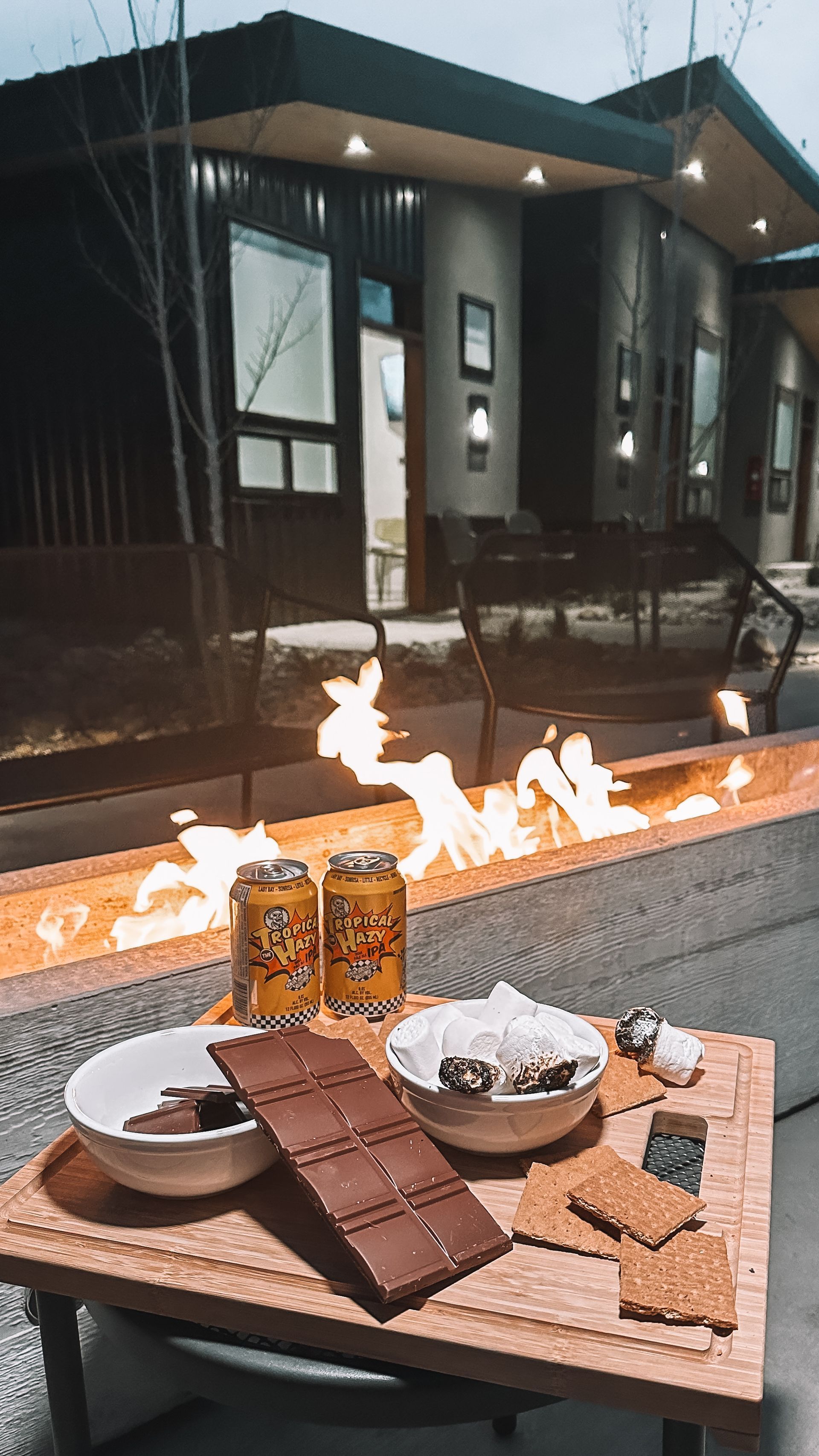 A s’mores kit with chocolate, graham crackers, and marshmallows sits on a table before an outdoor fire pit at dusk.