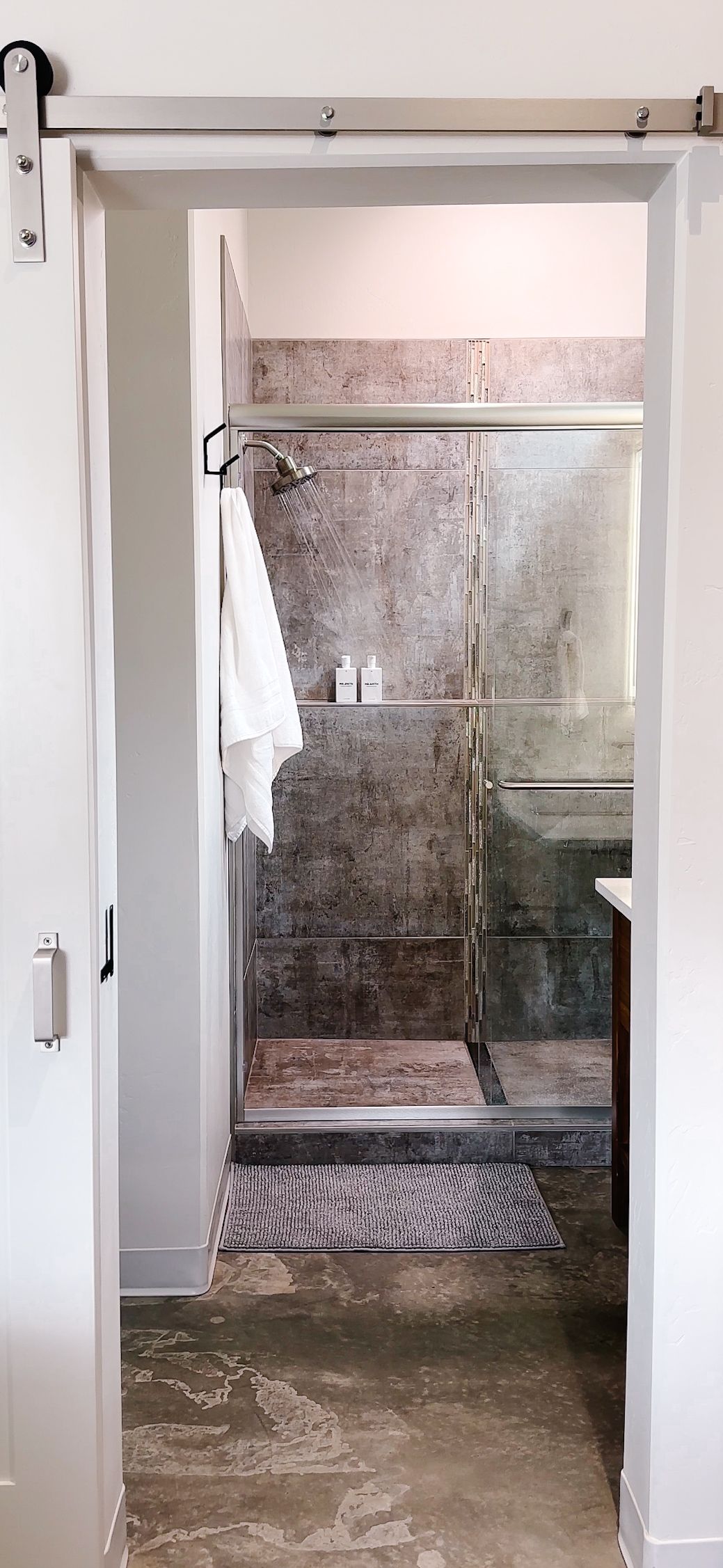 A bathroom shower with gray patterned tile and a glass door, viewed through an open white sliding barn door.