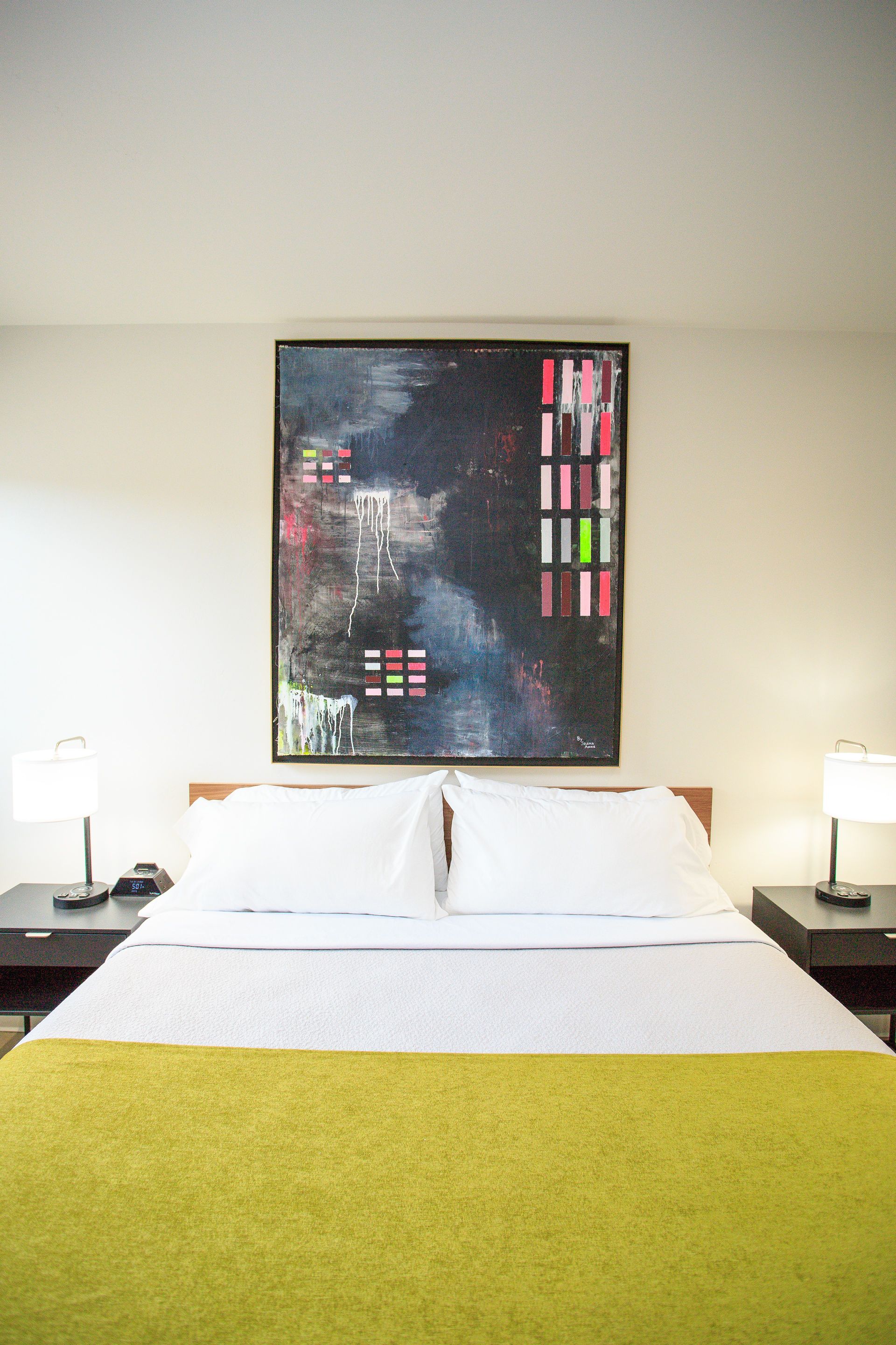 A bed with a bright yellow throw blanket, white pillows, and matching bedside lamps under an abstract dark-toned painting.