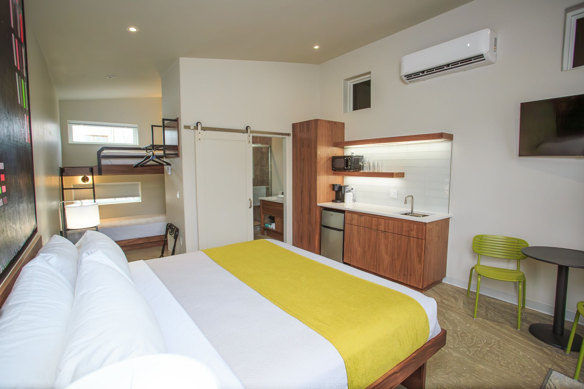 A hotel room with a queen bed, yellow runner, bunk beds, kitchenette, bathroom sliding door, and a table with green chair.