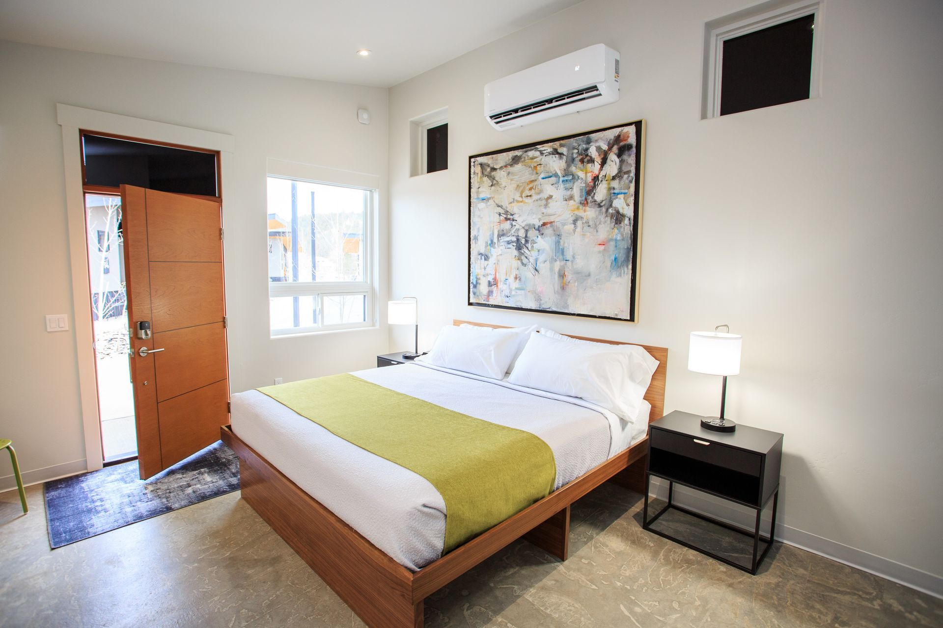 Modern bedroom with a wooden bed, lime green runner, large abstract painting, and an open door leading outside.