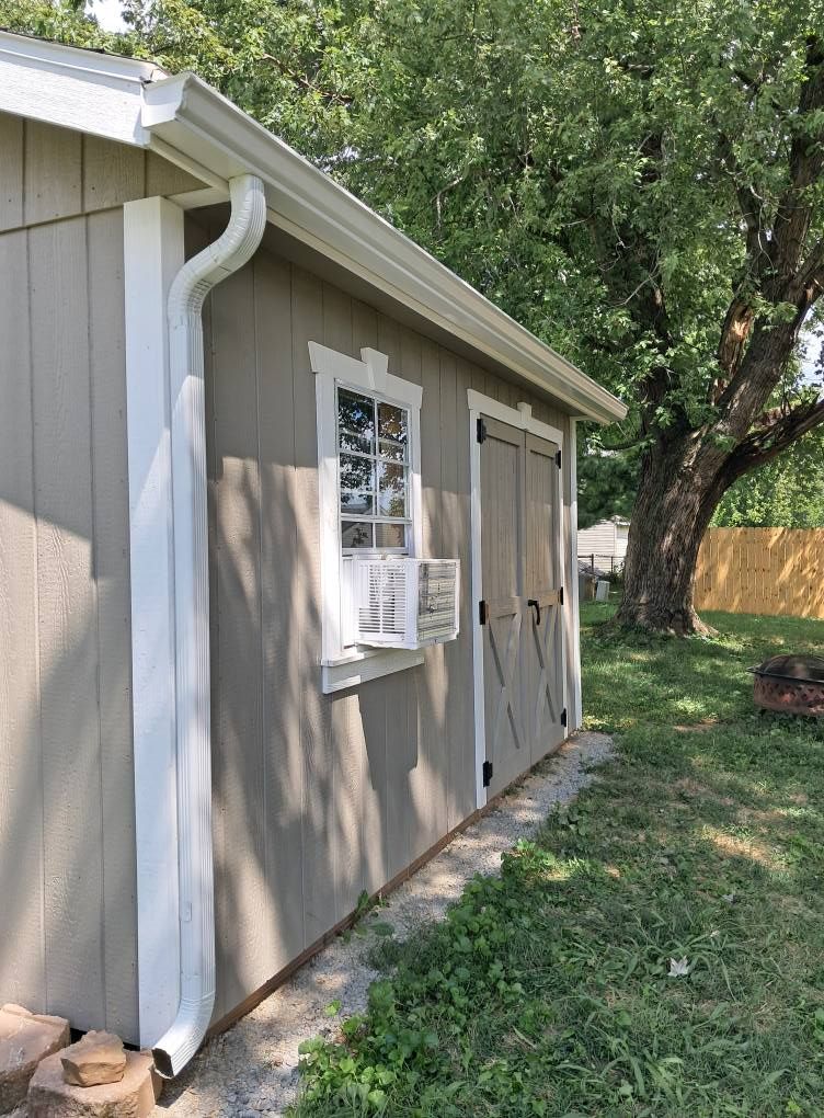 Tan shed with white trim, gutters, and a window with an AC unit. Next to a tree on grass.