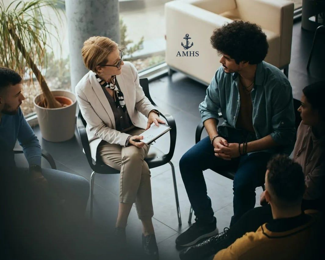 People seated in a circle during a discussion, with a woman speaking to a man in a modern room.