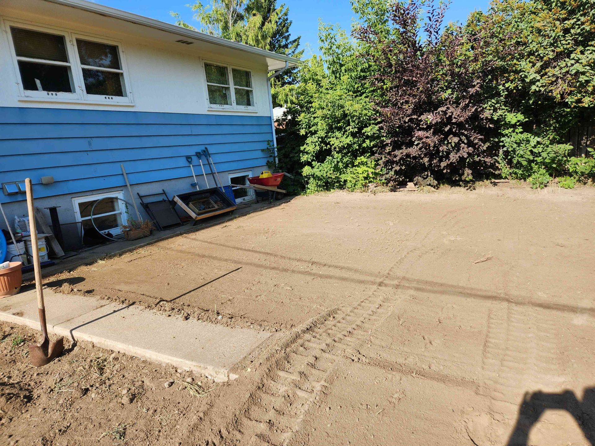 A house with blue siding borders a freshly graded, bare soil yard under a bright, clear blue sky.