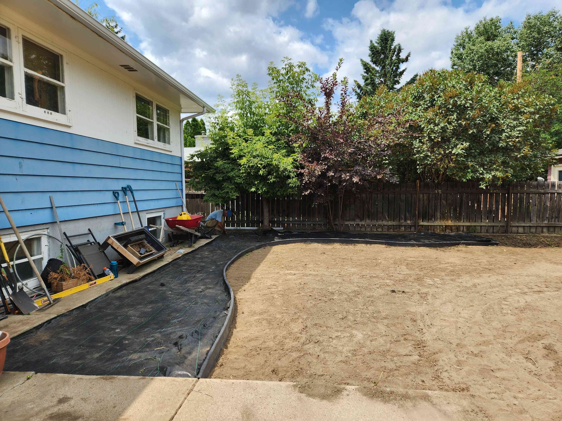A backyard landscape project showing a newly laid pathway base alongside a cleared dirt lawn area near a blue house.