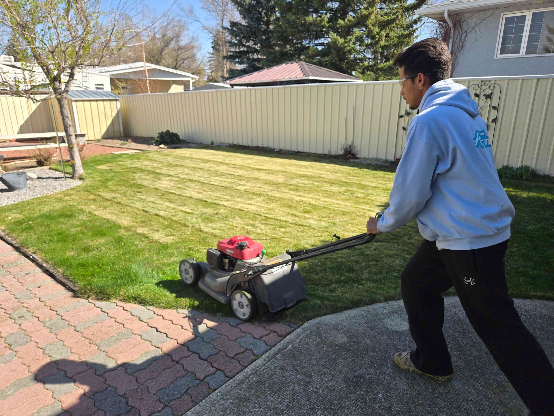 A person in a light blue hoodie mows a residential lawn with a grey walk-behind lawnmower on a sunny day.