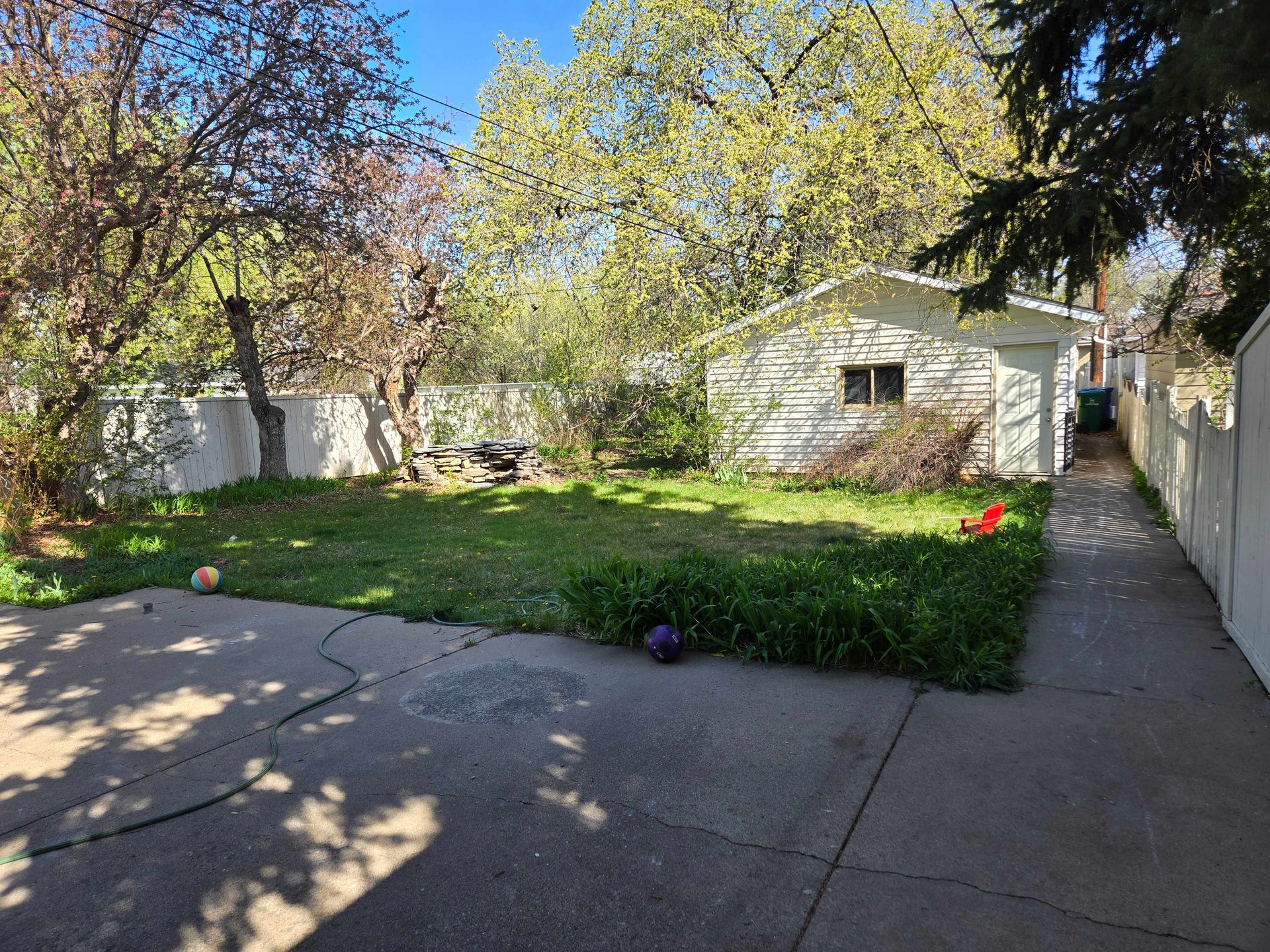 A backyard features a concrete patio in the foreground, a grassy lawn, trees, a white detached garage, and a side walkway.