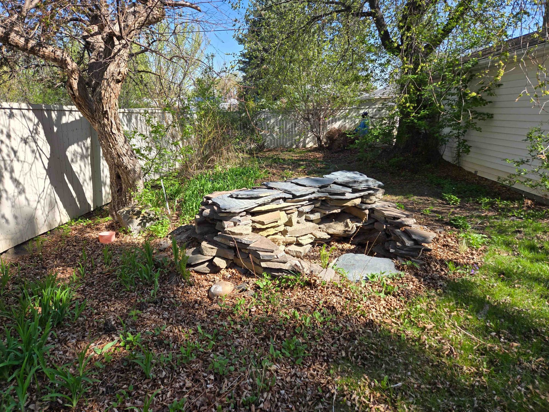 A stack of flat, layered rocks sits in a backyard garden with trees, grass, and a white fence.