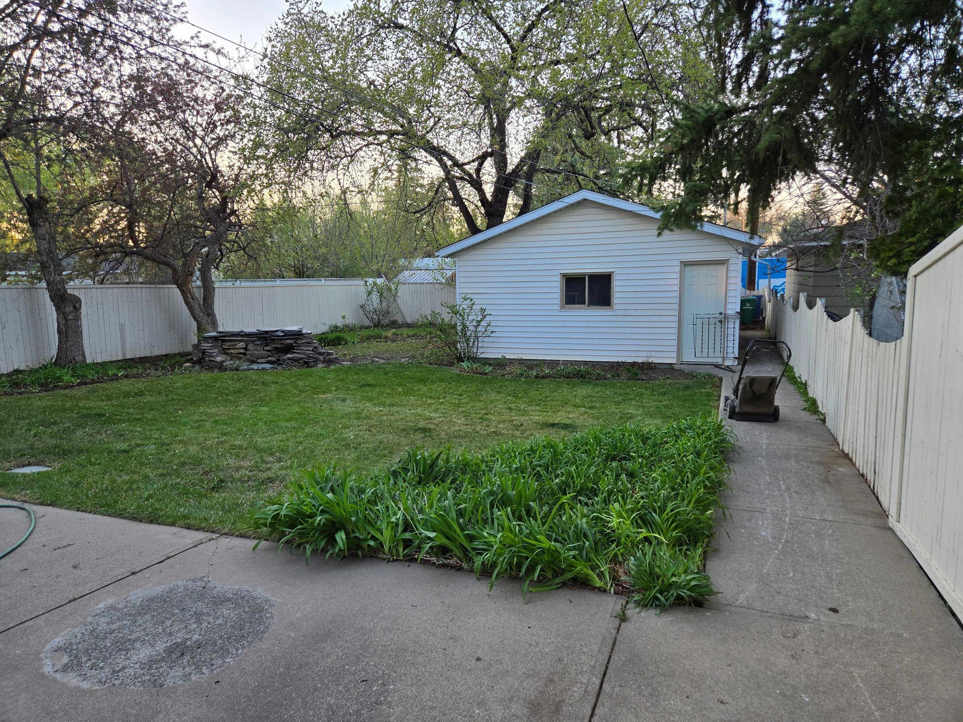 A backyard with a white detached garage, a small patch of green lawn, a garden bed, and a concrete path bordered by a fence.