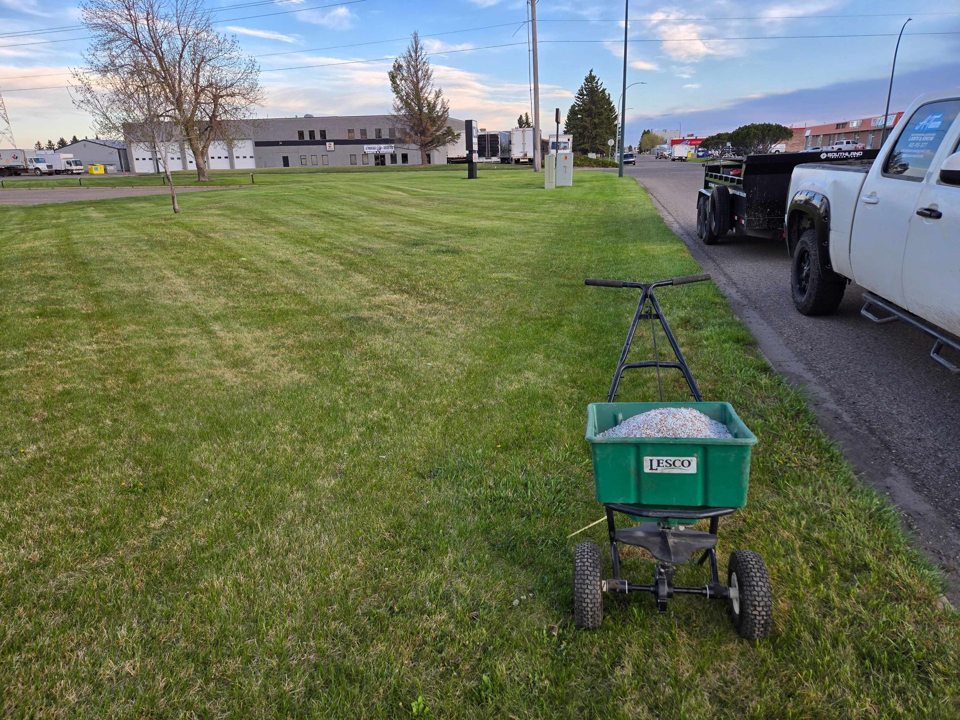 A green broadcast spreader sits on a grassy lawn next to a white pickup truck on an overcast day.