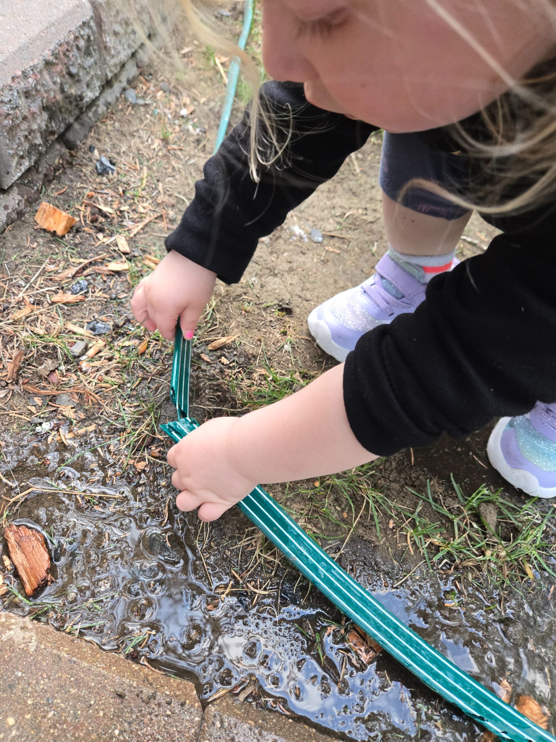 A person uses both hands to hold a green plastic edging strip down into wet, muddy soil near a brick pathway.
