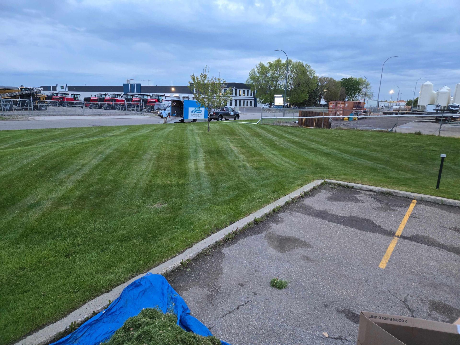 A freshly mowed green lawn with distinct mowing lines, seen from a paved parking lot under a cloudy sky.