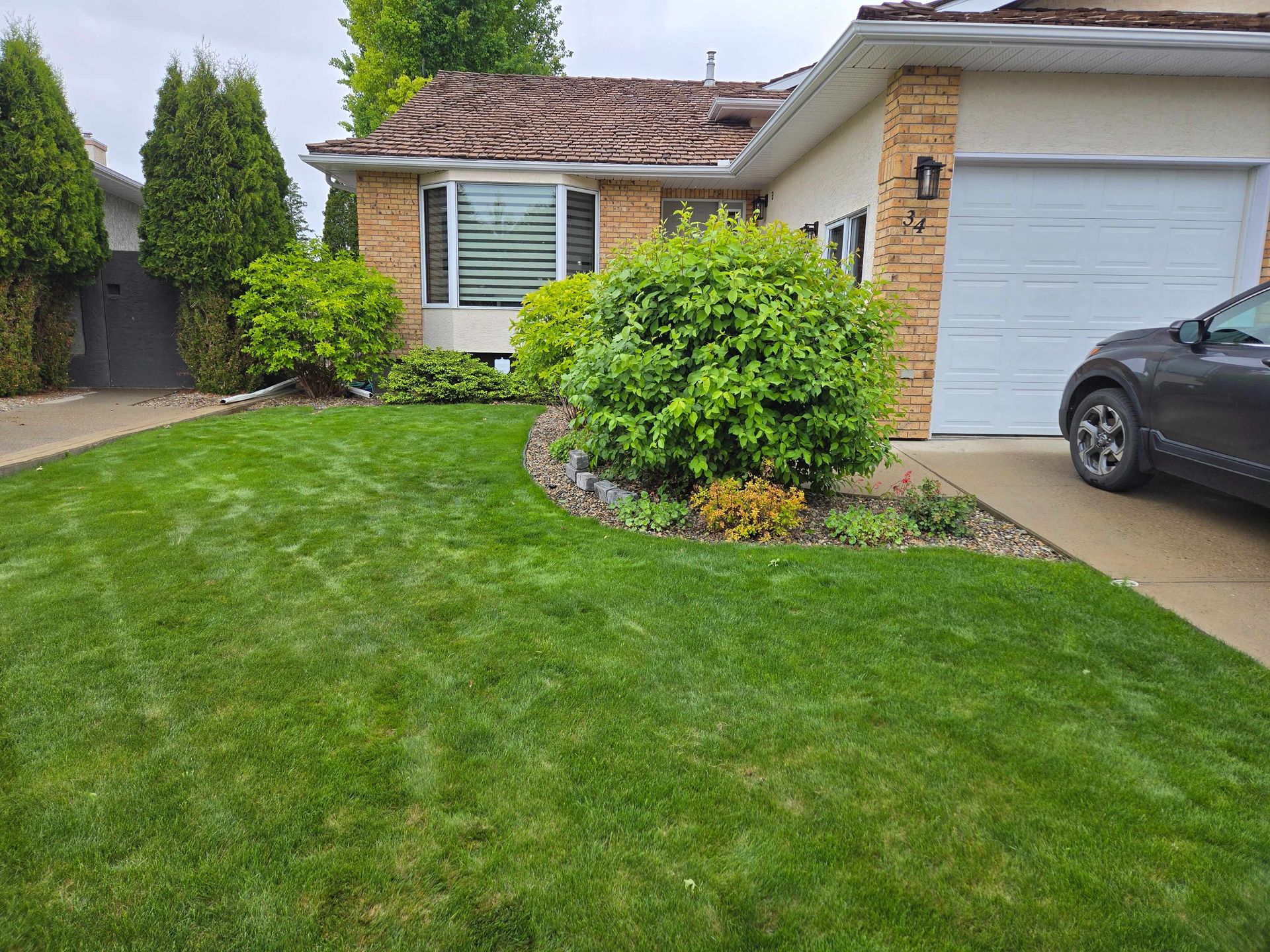 A suburban house with a tan brick exterior, a grey garage door, green lawn, and landscaped shrubs in the front yard.