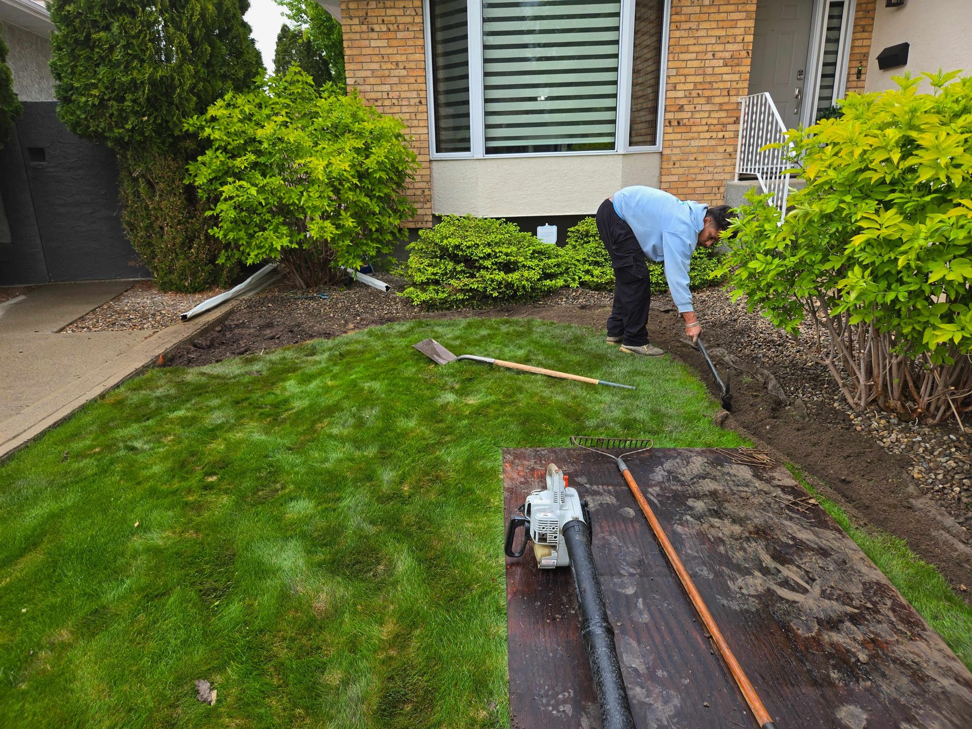 A person in a light blue shirt trims the edge of a residential lawn next to a garden bed. A leaf blower sits nearby.