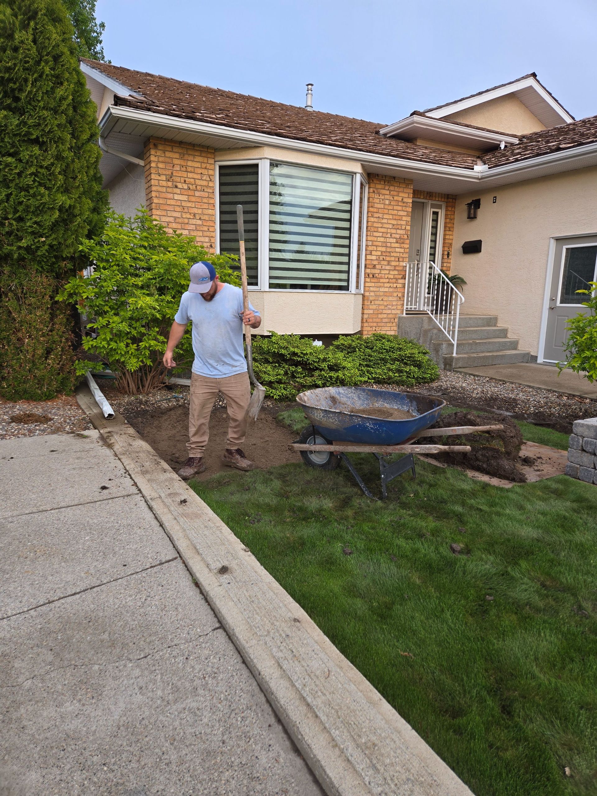 A worker in a light blue shirt and tan pants stands next to a wheelbarrow while digging in a yard near a house.