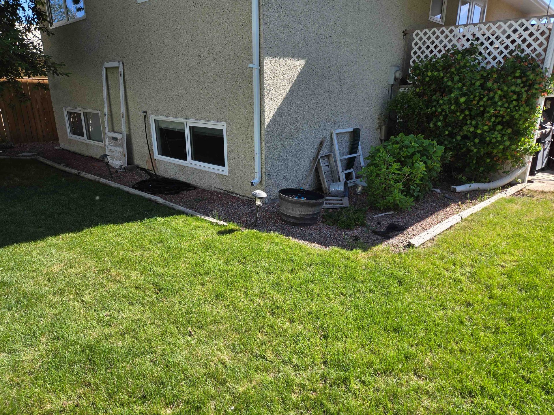 Stucco house side wall with windows, a lawn, and a garden bed filled with dark mulch and a green bush.