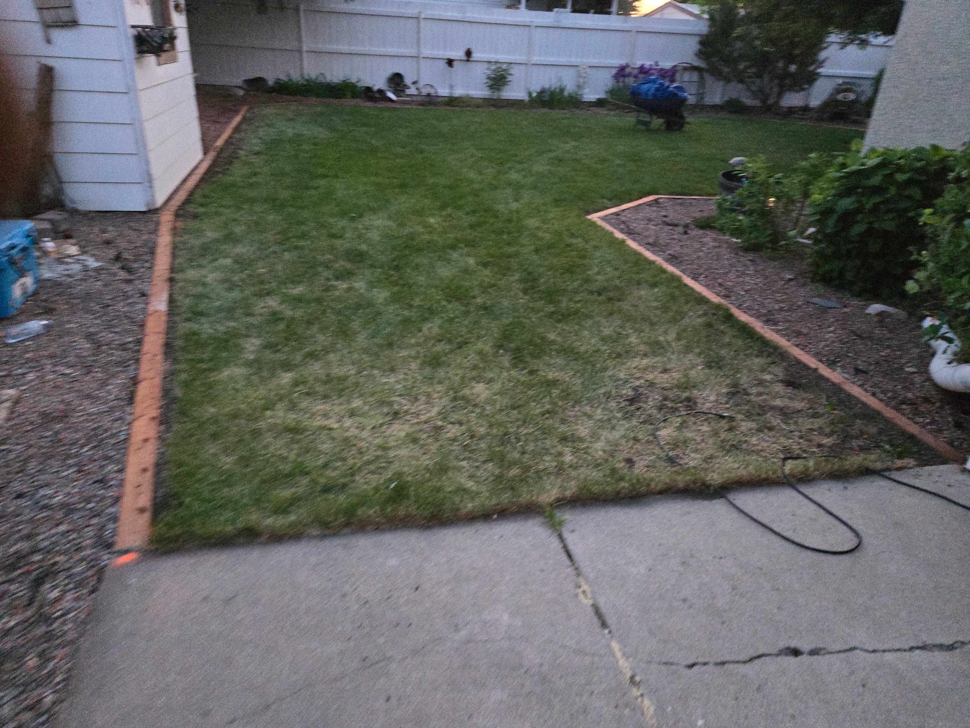 A backyard lawn with concrete patio in the foreground, framed by brick edging and a gravel border.