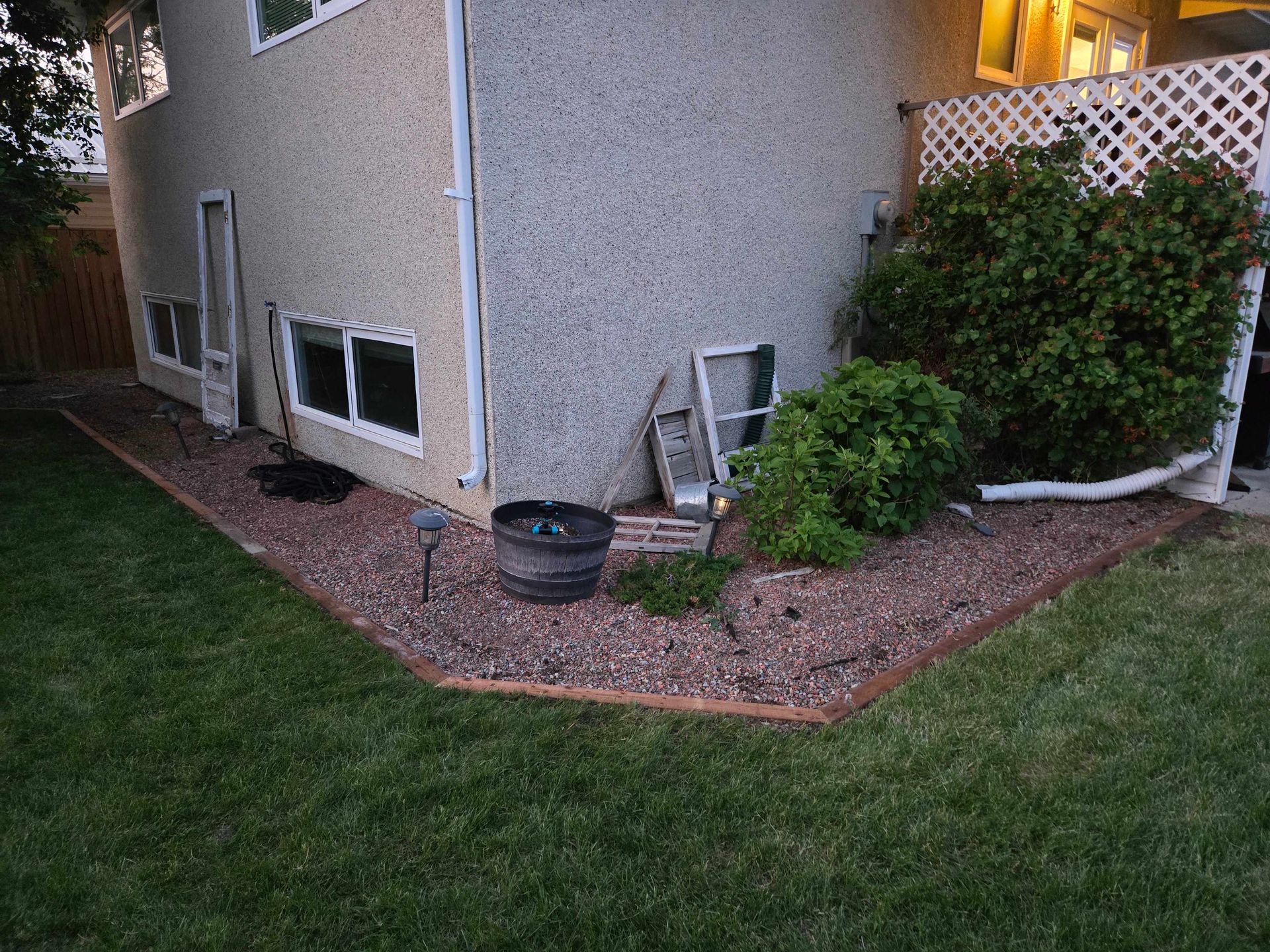 A house exterior corner with a gravel garden bed, a wooden border, scattered window frames, and a green shrub.