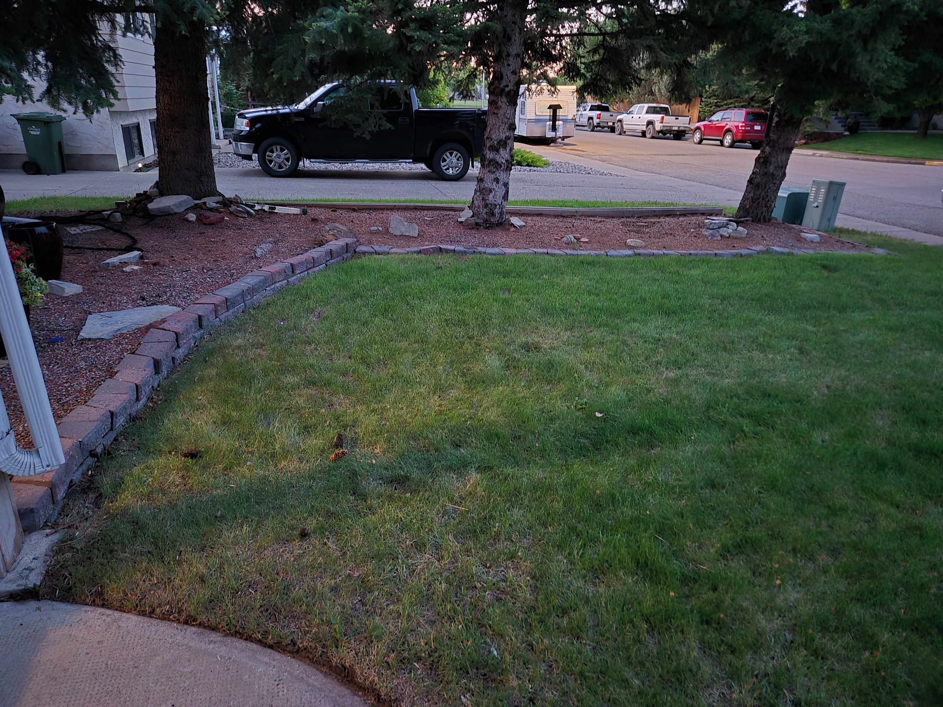 A residential front yard at dusk with green grass, a red rock garden bed, brick edging, and parked cars on the street.