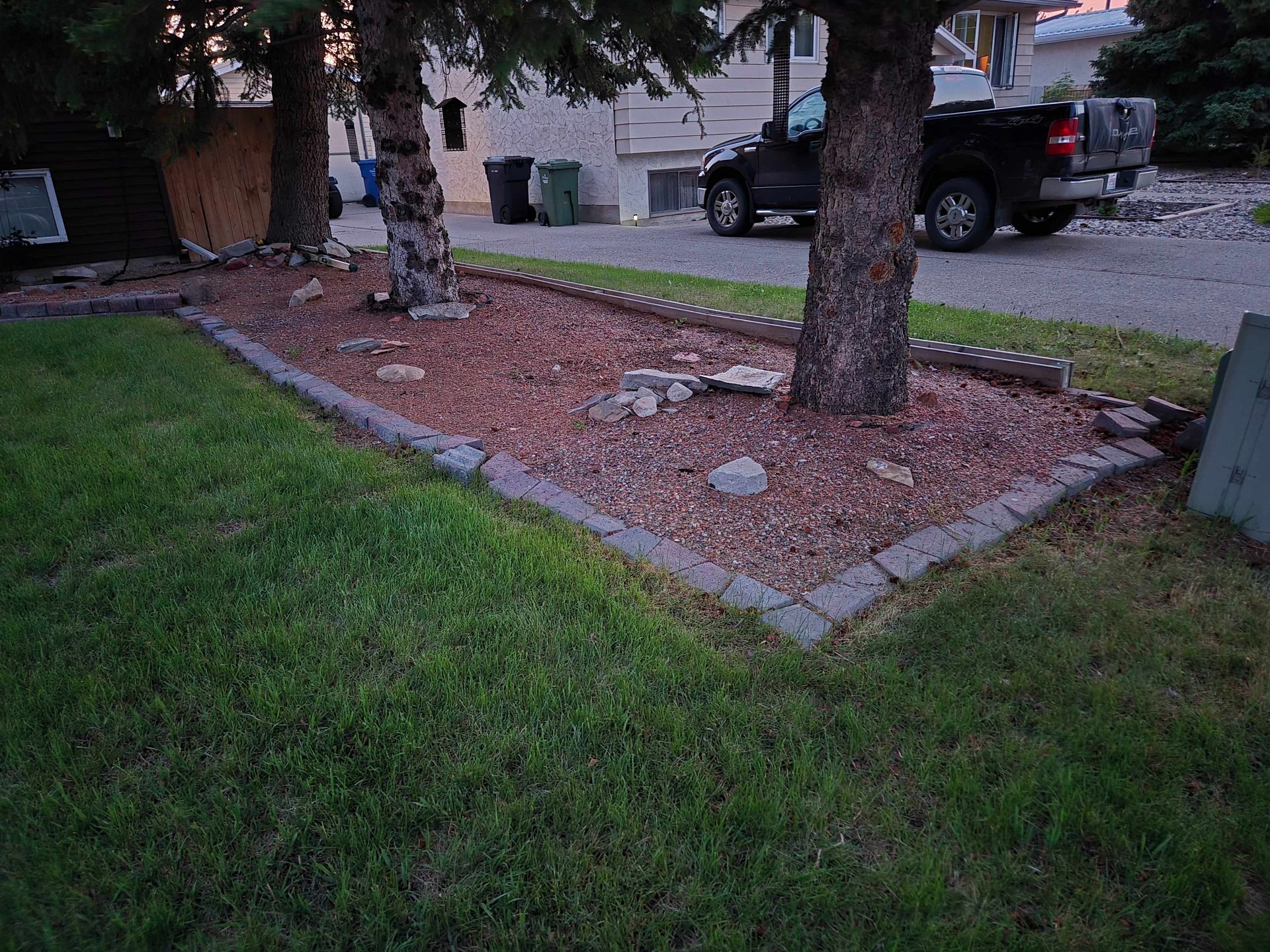A landscaped yard featuring two trees, a red mulch bed bordered by grey pavers, a green lawn, and a parked black truck.