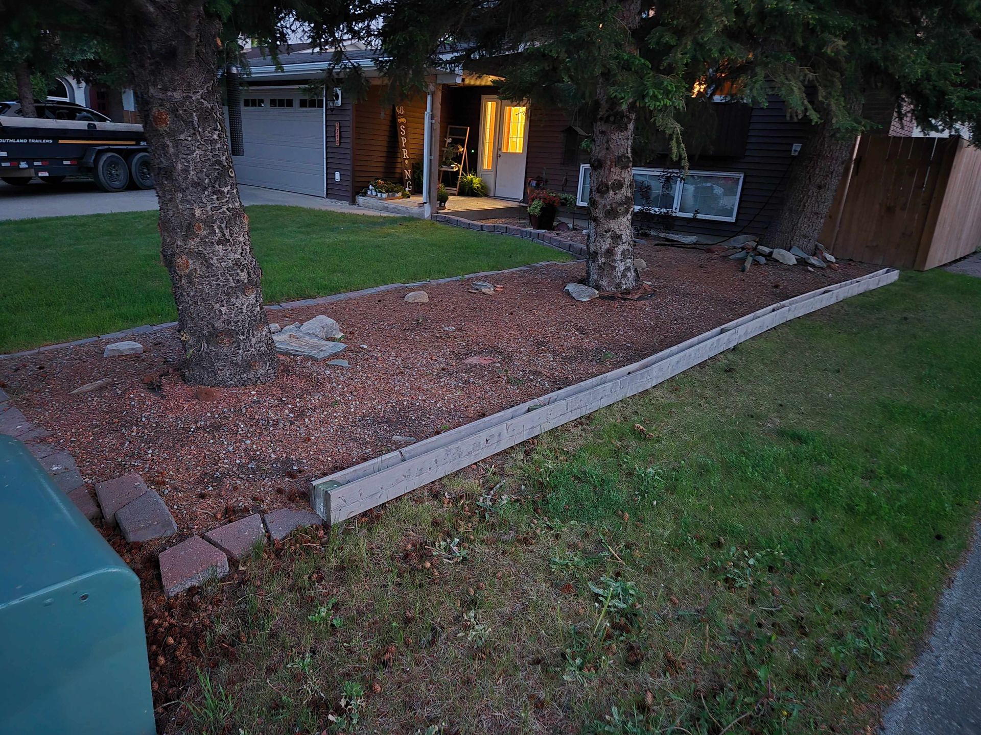 A front yard garden bed with reddish mulch, two mature evergreen trees, and a concrete block retaining wall border.