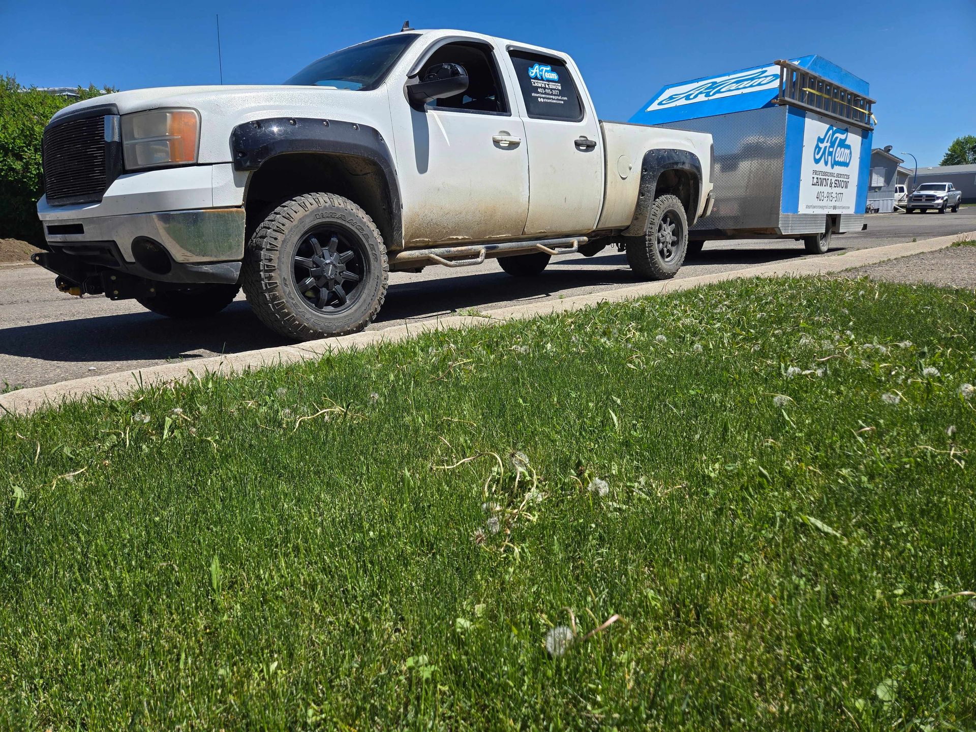 A white pickup truck with black wheels parked on the side of a road, towing a small, blue and silver enclosed trailer.