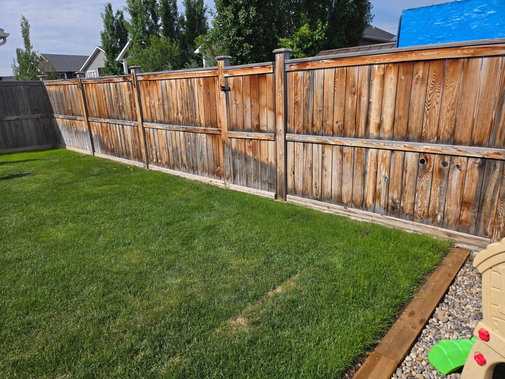 A view of a backyard featuring a weathered wooden fence with a grass lawn and a gravel border with landscaping timber.