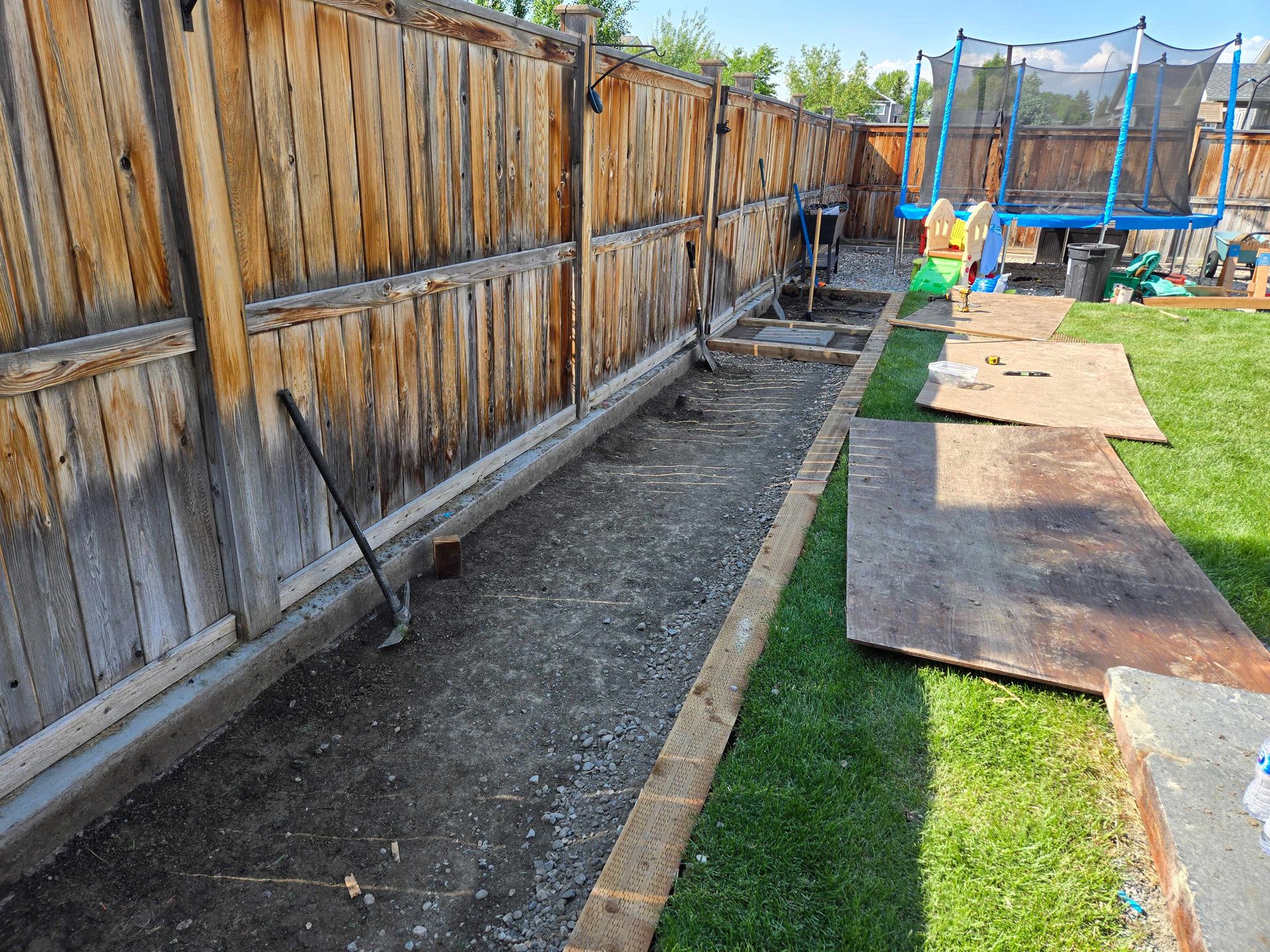 A backyard side yard features a dirt trench lined with wooden beams, running alongside a weathered wooden fence.