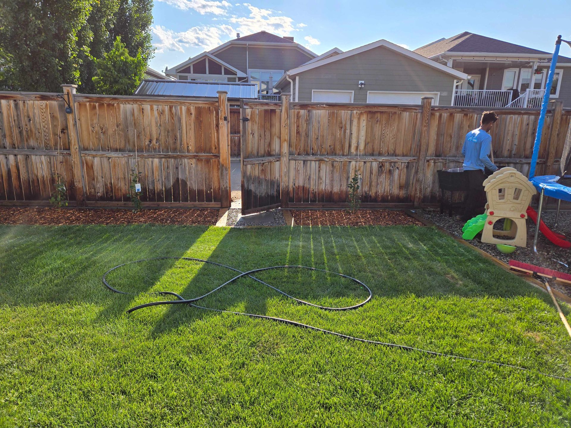 A sunlit backyard with a green lawn, a wooden fence, a garden hose, and a child’s plastic playhouse in the corner.