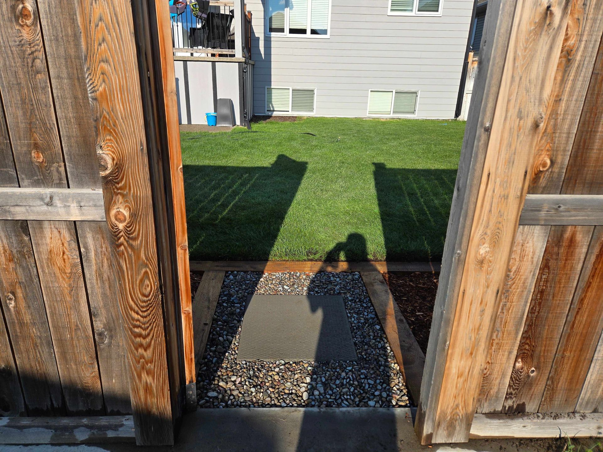 A wooden fence gate opens onto a gravel path leading to a backyard with green grass and a house exterior.
