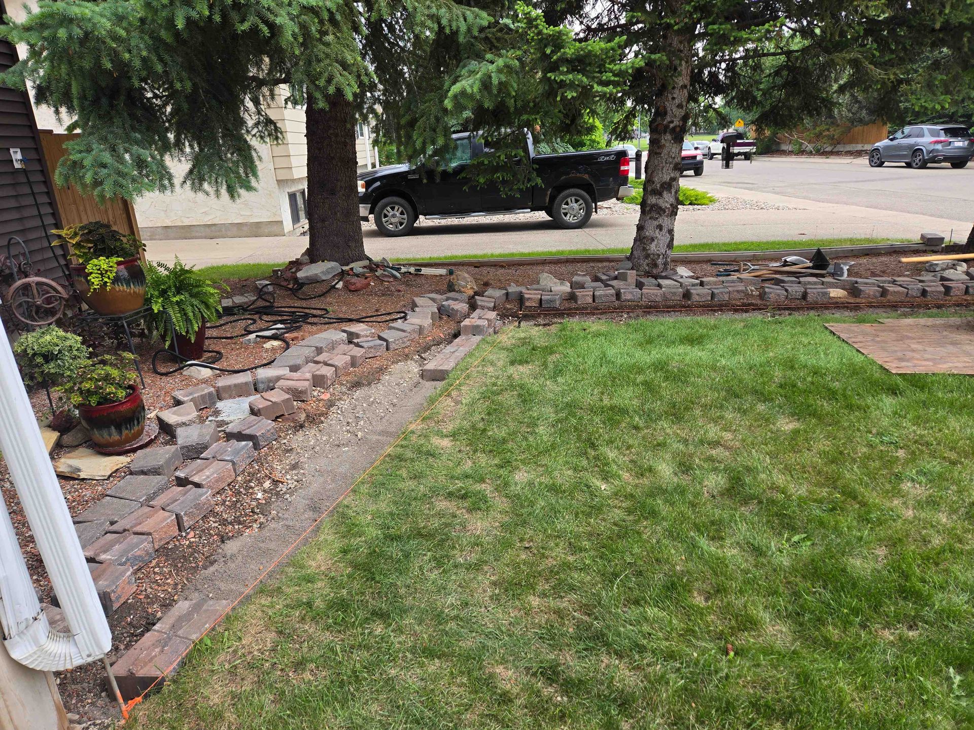 A lawn with a stone-bordered garden bed featuring mulch, small shrubs, and potted plants, near a house and street.